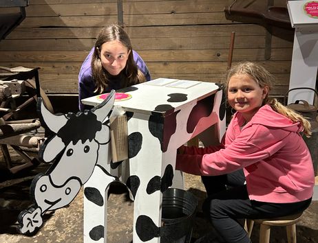 Zwei Ferien-Kinder sitzen auf dem Melkschemel und melken die Holzkuh 