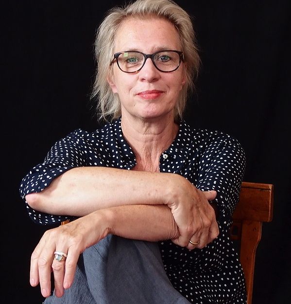 Frau mit Brille und gepunkteter Bluse sitzt auf einem Stuhl vor schwarzem Hintergrund.