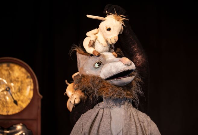 Eine Puppenfigur eines Wolfes mit einem kleinen Ziegenbock auf dem Kopf, vor einem dunklen Hintergrund und einer Uhr.