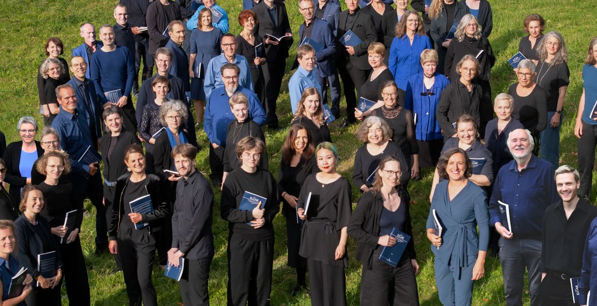 Eine Gruppe von Menschen steht auf einer Wiese und hält Notenblätter in den Händen.