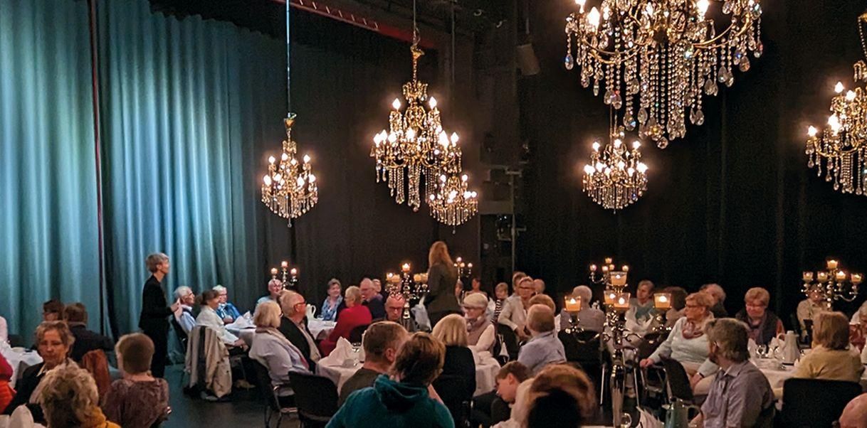 Menschen sitzen an gedeckten Tischen in einem Theatersaal mit Kronleuchtern.