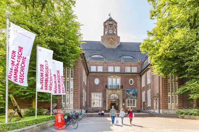 Menschen vor einem palastähnlichen Gebäude. Der rote Backsteinbau geht über 5 Etagen. Der Eingangsbereich ist über Treppen erreichbar, hat ein steinernes Tor und ist von Fenstern umrandet. Darüber ein Balkon. Auf dem Dach ist mittig ein Turm.