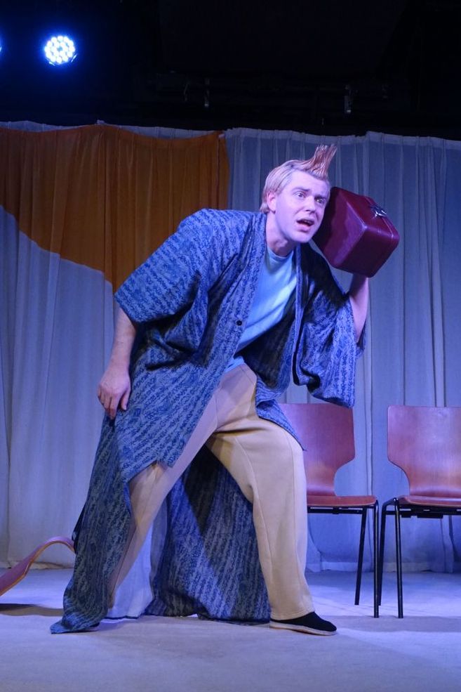 Ein Schauspieler in einem blauen Kimono und beigen Hosen hält einen roten Koffer ans Ohr auf einer Bühne.