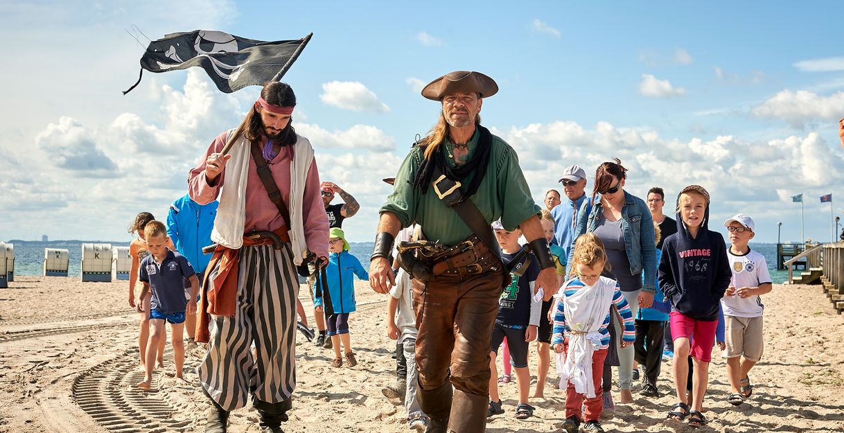 Eine Gruppe von Menschen, darunter zwei als Piraten verkleidete Männer, spaziert am Strand entlang. Kinder und Erwachsene folgen ihnen. Der Himmel ist blau mit Wolken.