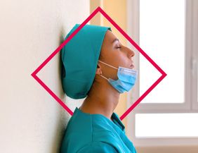 Eine Frau mit dem Kopf angelehnt an eine Wand mit einer grünen Haube und einem Mundschutz vor dem Kinn hat die Augen geschlossen.