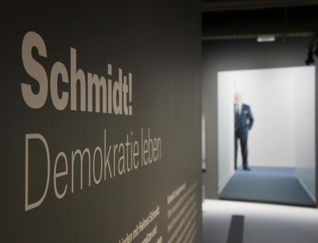 Fotos, das eine von der Seite aufgenommene Informationstafel im Vordergrund zeigt. Darauf ist deutlich "Schmidt! Demokratie leben" zu lesen. Im Hintergrund steht hinter einem Durchgang ein Mann im Anzug, der nur verschwommen zu sehen ist.