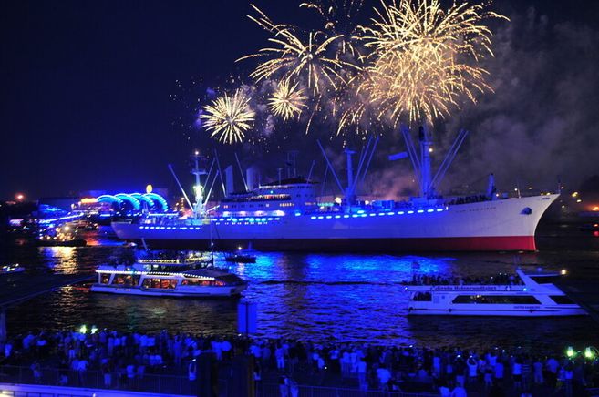 Ein beleuchtetes Schiff mit Feuerwerk am Nachthimmel in einem Hafen.