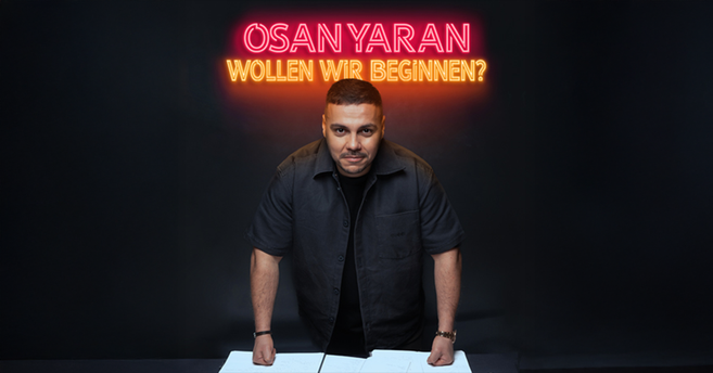 Ein Mann steht vor einem Tisch mit Neon-Schriftzug 'Osan Yaran - Wollen wir beginnen?' im Hintergrund.
