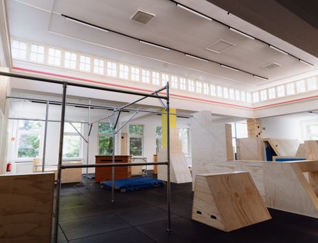 Zu sehen ist, die menschenleere Parkourhalle mit sanftem Morgenlicht durch die Oberlichter. Hindernisse für das Training aus Holz und Metal stehen bereit.