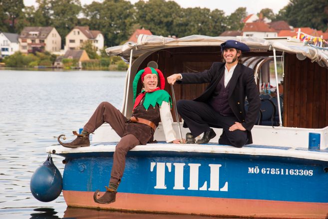 Zwei Männer in historischen Kostümen sitzen auf einem Boot mit dem Namen 'TILL'.