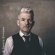 Porträt eines Mannes mit weißem Haar, Anzugweste und Krawatte vor einem neutralen Hintergrund.