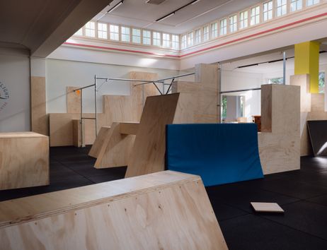 In der hellen Parkourhalle mit Oberlichtern und dunklem Gummi Boden finden sich unterschiedlich Holzhindernisse und Stangen für das Parkourtraining.