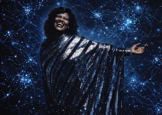 Eine Frau steht in einem silber-glitzernden Kleid vor dem Hintergrund einer dunkelblauen Fläche, auf der viele Lichter sind.