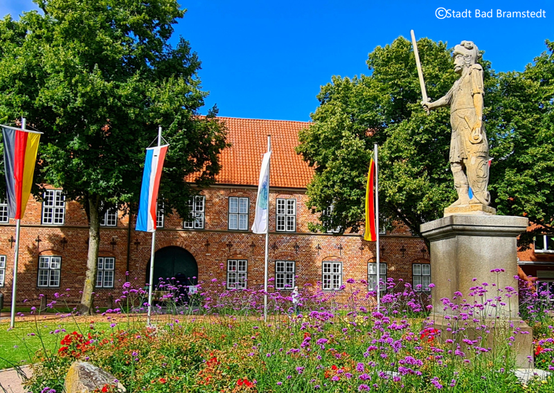 Statue vor einem historischen Gebäude mit Flaggen und Blumenbeet in Bad Bramstedt.