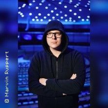 Person in schwarzem Hoodie mit verschränkten Armen vor einer Bühne mit blauer Beleuchtung.