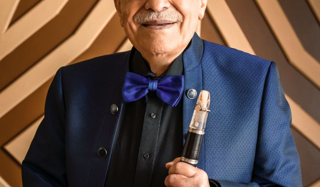 Ein älterer Mann in einem blauen Anzug mit Fliege hält eine Klarinette vor einem geometrischen Hintergrund.