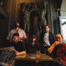 Fünf Männer sitzen an einem Tisch in einem stilvollen Raum mit antiken Statuen im Hintergrund.