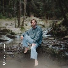 Eine Person sitzt barfuß auf einem Felsen in einem bewaldeten Flussbett.