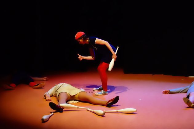 Eine Person in roter Kleidung steht auf einer Bühne mit Jonglierkeulen, umgeben von drei liegenden Personen.