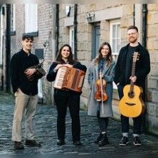 Vier junge Musiker mit Instrumenten stehen auf einer gepflasterten Straße vor einem Gebäude.