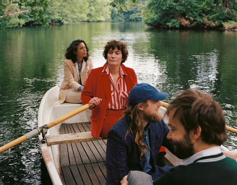 Die Band sitzt in einem Ruderboot auf einem  See oder Fluß, zwei Frauen im Bug, zwei Männer im Heck, Sängerin/Frontfrau im roten Anzug rudert