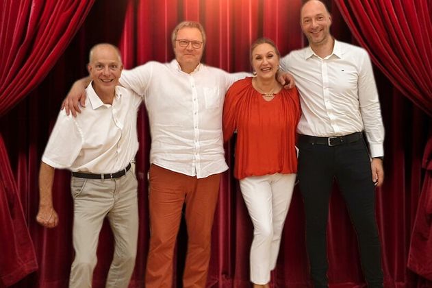 Vier Personen stehen vor einem roten Bühnenvorhang und lächeln in die Kamera.