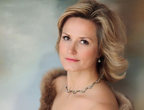 Porträt einer Frau mit blondem Haar und elegantem Schmuck vor einem unscharfen Hintergrund.