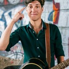 Ein Mann mit Gitarre zeigt auf seinen Kopf vor einer Graffiti-Wand.