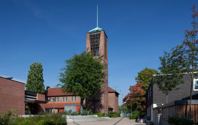 Eine Kirche mit einem hohen Turm und rotem Ziegeldach, umgeben von Bäumen und einem klaren blauen Himmel.