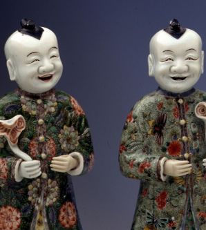 Zwei freundlich lächelnde Porzellanfiguren. Sie wirken männlich, ihr Gewand besteht aus Rock und Mantel, verziert mit chinesischen Mustern. Beide haben glattrasierte Köpfe mit einem Haarstreifen in der Mitte und halten etwas in der Hand.