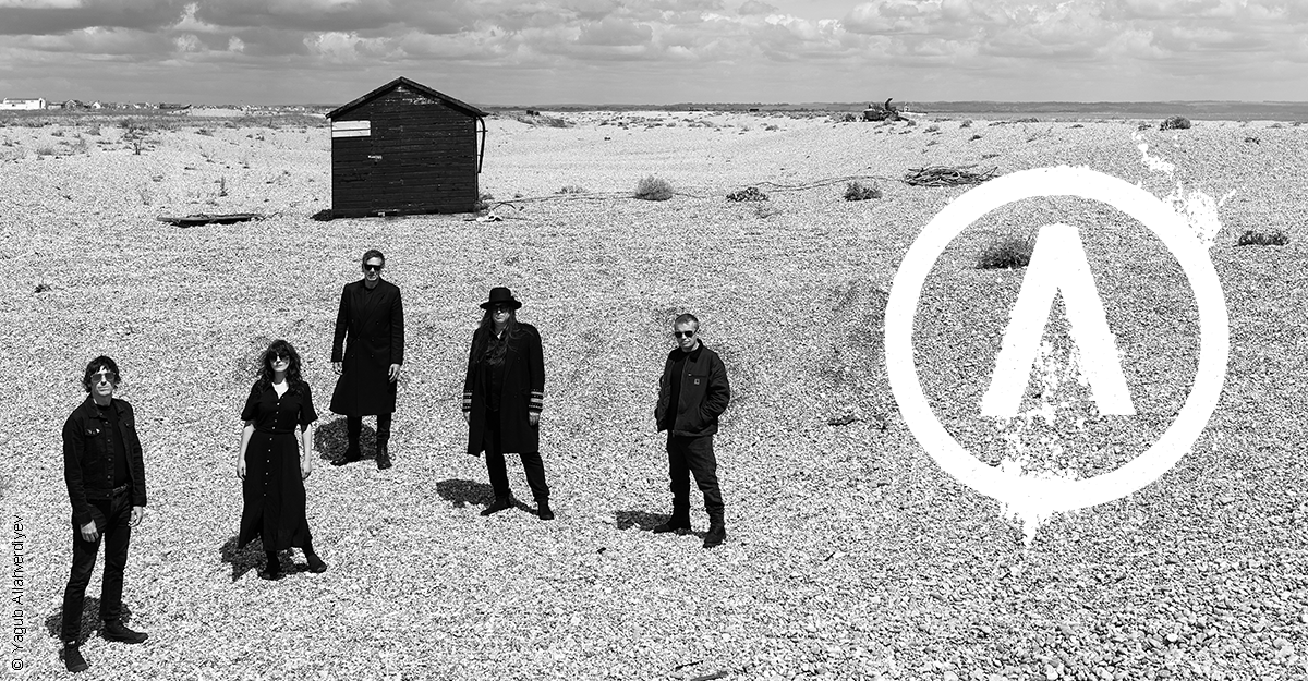 Fünf Personen stehen auf einem Kiesstrand vor einer Holzhütte, mit einem großen weißen Logo im Vordergrund.