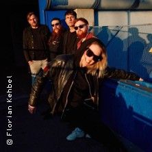 Gruppe von fünf Personen in lässiger Kleidung und Sonnenbrillen posiert vor einer blauen Wand.