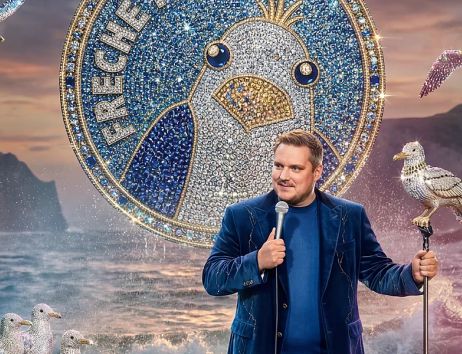 Ein Mann hält ein Mikrofon und steht vor einem großen, glitzernden Möwen-Logo. Im Hintergrund sind das Meer und Klippen zu sehen.