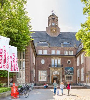 Menschen vor einem palastähnlichen Gebäude. Der rote Backsteinbau geht über 5 Etagen. Der Eingangsbereich ist über Treppen erreichbar, hat ein steinernes Tor und ist von Fenstern umrandet. Darüber ein Balkon. Auf dem Dach ist mittig ein Turm.