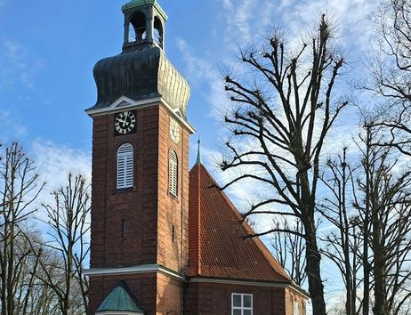 Backsteinkirche mit grünem Turm und Uhr, umgeben von kahlen Bäumen, unter blauem Himmel.