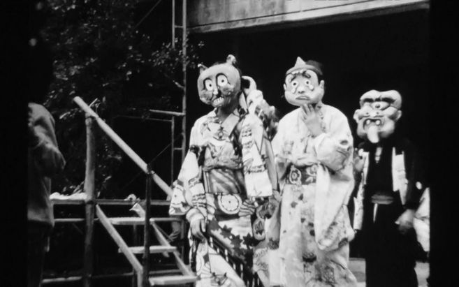 Schwarz-Weiß-Foto von Personen in traditionellen Kostümen und Masken auf einer Bühne.