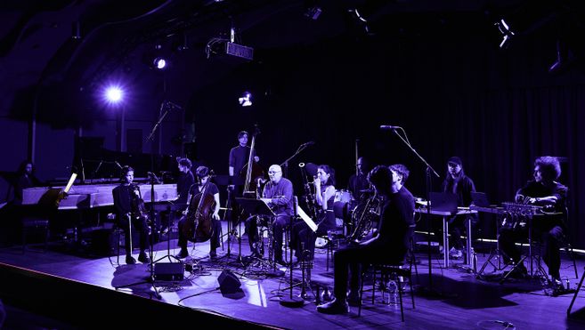 Ein Ensemble von Musikern auf einer Bühne mit verschiedenen Instrumenten, beleuchtet von violettem Licht.