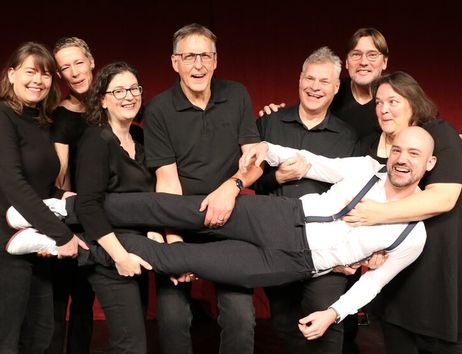 Eine Gruppe von acht Personen in schwarzer Kleidung hält lachend einen Mann in weißem Hemd und Hosenträgern horizontal vor einem roten Hintergrund.