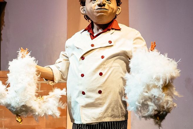 Ein Schauspieler in einem weißen Kochkostüm mit einer großen Maske hält zwei Hühner aus Federn in den Händen.