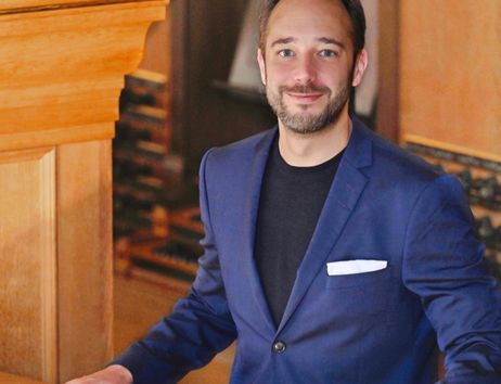 Ein Mann in einem blauen Anzug steht an einer Holzbalustrade und lächelt in die Kamera.