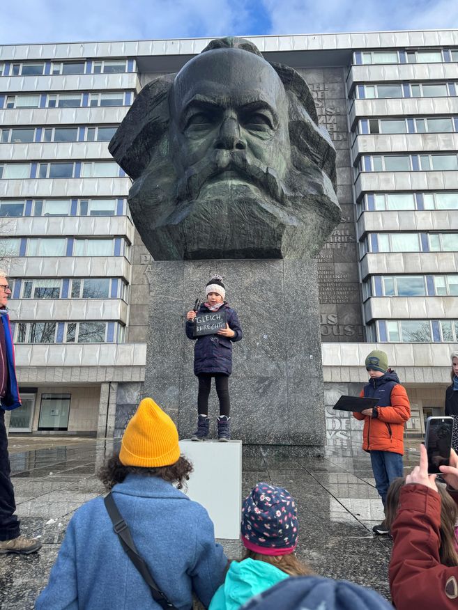 A Kid stands on a base in front of a huge statue of Karl Marx.