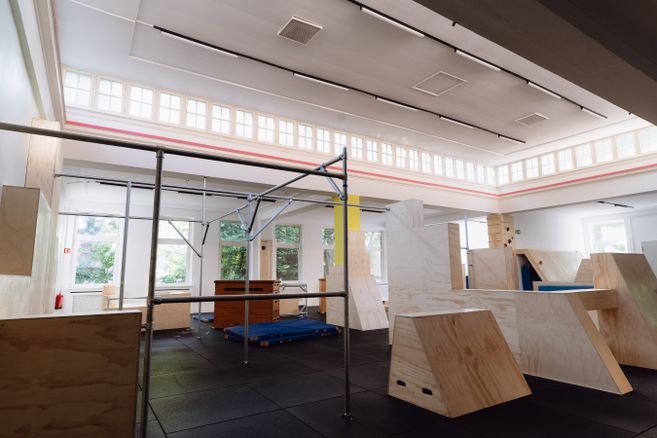 Zu sehen ist, die menschenleere Parkourhalle mit sanftem Morgenlicht durch die Oberlichter. Hindernisse für das Training aus Holz und Metal stehen bereit.