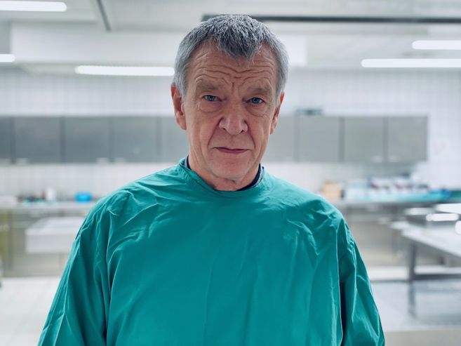Ein älterer Mann in einem grünen Kittel steht in einem sterilen Raum, möglicherweise einem Labor oder einer Pathologie.