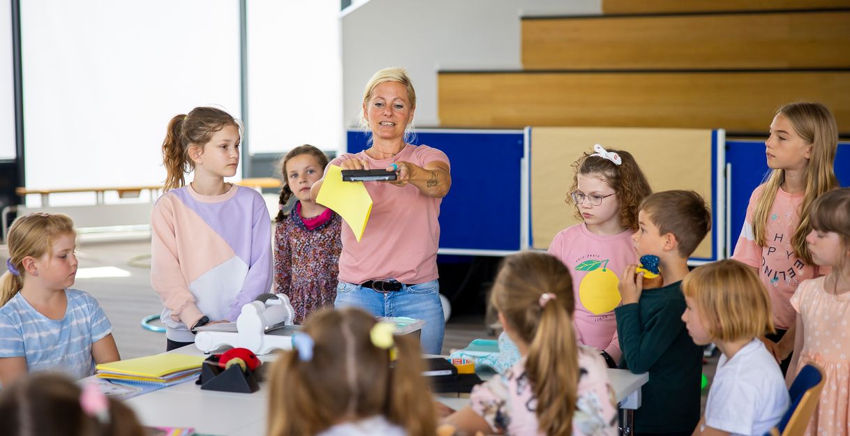Eine Frau zeigt einer Gruppe von Kindern ein gelbes Blatt Papier in einem Klassenzimmer.