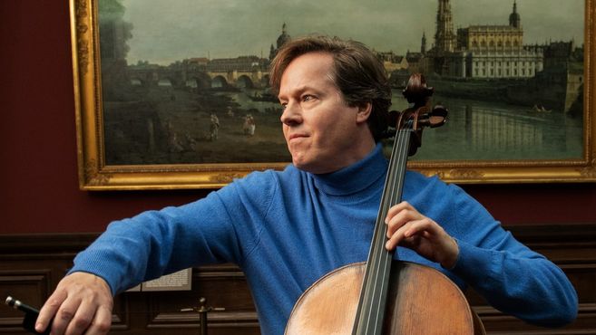 Ein Mann in einem blauen Pullover spielt Cello vor einem Gemälde in einem Museum.