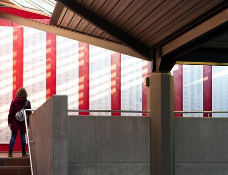Eine Person betrachtet eine Wand mit langen Listen von Namen, die auf weißen Tafeln vor einem roten Hintergrund angebracht sind.