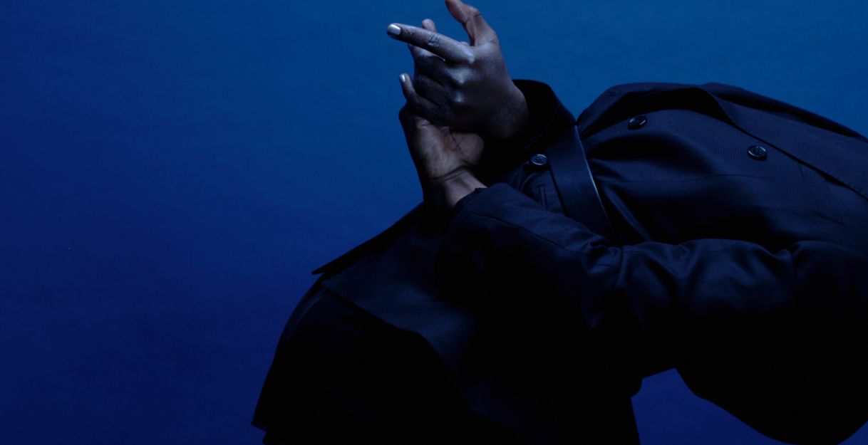 A performer is seen from the side, leaning forward, a black hoodie pulled over their head. Their hands are behind their back and stretched out.