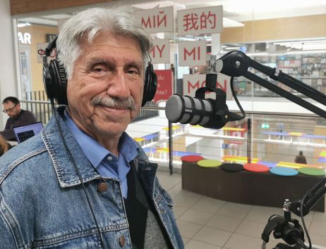 Ein älterer Mann mit Kopfhörern steht vor einem Mikrofon in einem Café oder Studio.