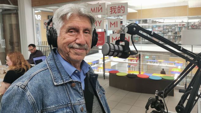 Ein älterer Mann mit Kopfhörern steht vor einem Mikrofon in einem Café oder Studio.