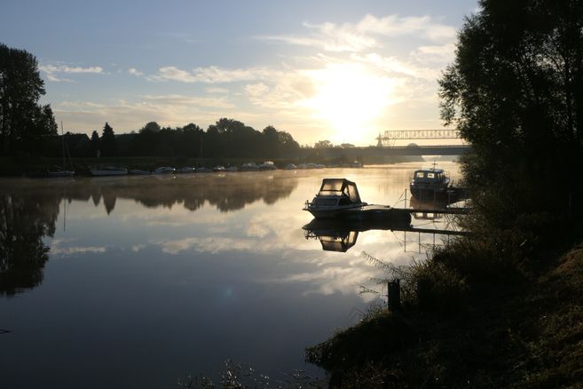 Boote auf einem ruhigen Fluss bei Sonnenaufgang mit Brücke im Hintergrund.